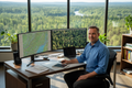 Professional forestry consultant at desk with dual monitors showing forest maps, home office with forest view through window